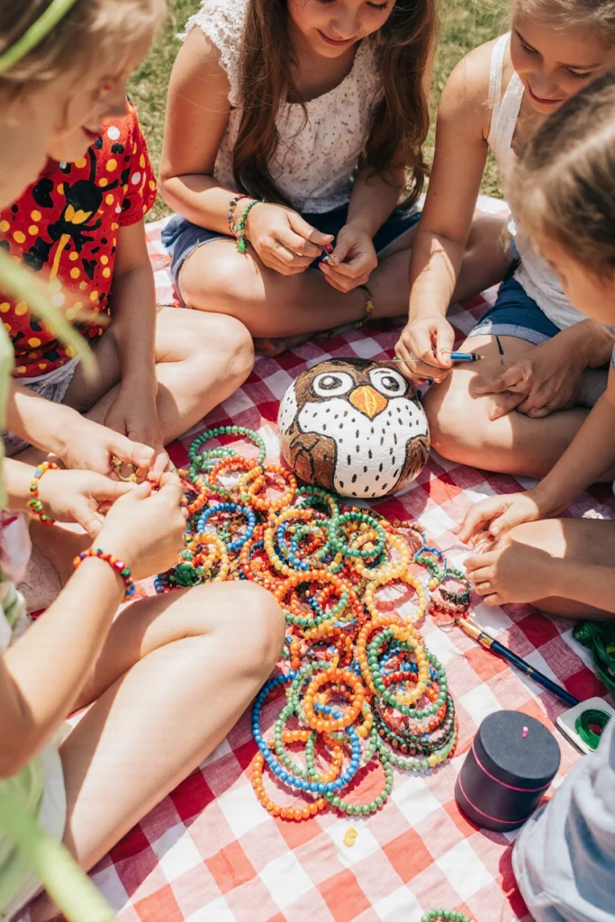 Enfants assis sur un plaid vichy rouge en plein air, fabriquant des bracelets d’amitié en perles multicolores autour d’un gros galet peint en chouette.